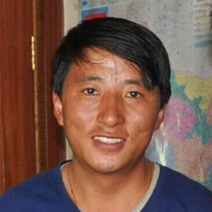 A smiling person with short black hair wearing a blue shirt stands indoors in front of a map on the wall.