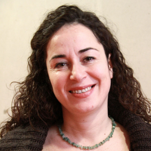 A woman with curly dark hair is smiling at the camera. She is wearing a brown sweater and a green beaded necklace, standing against a plain light-colored background.