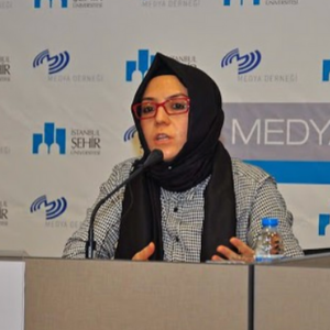 A woman wearing a black headscarf and glasses speaks at a conference table with a microphone. A water bottle is in front of her, and a backdrop displays logos for Istanbul Şehir University and Medya Derneği.