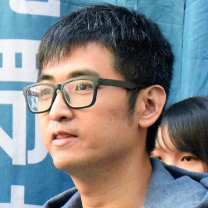 A man with short black hair and glasses stands in front of a blue background with white text. A woman with long dark hair is partially visible behind him.