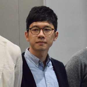 A young man with short black hair and glasses wearing a light blue collared shirt and a dark sweater stands indoors, looking slightly to his left.