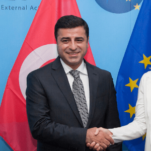 A man in a dark suit and patterned tie smiles while shaking hands, standing in front of Turkish and European Union flags.