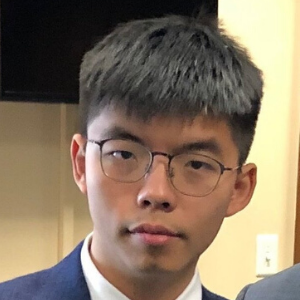 A young person with short black hair and glasses, wearing a dark suit and white shirt, stands indoors in front of a beige wall.