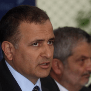 A man in a suit speaking, with another man in the background, both in focus. The setting appears formal, possibly a meeting or press conference.
