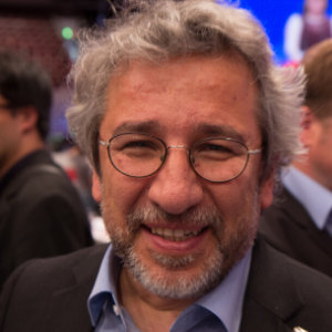 A smiling man with gray curly hair, glasses, and a beard, wearing a dark suit and light blue shirt, stands in a crowded indoor setting. The background is blurred with people and a blue screen visible.