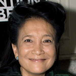 A woman with dark hair styled back, wearing a green top and blue earrings, smiles at the camera against a blurred background with partial black and white text.