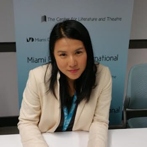 A woman with long black hair, wearing a cream-colored blazer and blue lanyard, sits at a table in front of a blue banner displaying The Center for Literature and Theatre and Miami Book Fair International.