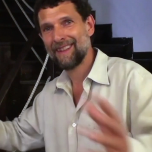 A man with short curly hair and a beard, wearing a light-colored shirt, smiles and gestures with his hands indoors near a staircase with rope railing.