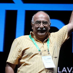 An older man with a mustache, glasses, and a yellow striped shirt smiles and raises his arm on stage. He is wearing a green lanyard and name badge. The background is dark with large blue letters partially visible.