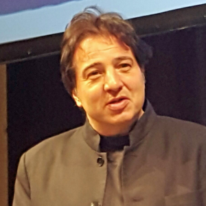 A man with dark hair wearing a dark-colored jacket is speaking or presenting in front of a dark background and a projected screen.