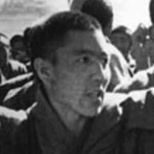 A black and white photo showing a man with short hair in a crowd, looking to the side, with several other people visible in the blurred background.