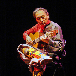 A person in colorful clothing and boots sits on a chair, playing an acoustic guitar under stage lighting against a dark background.