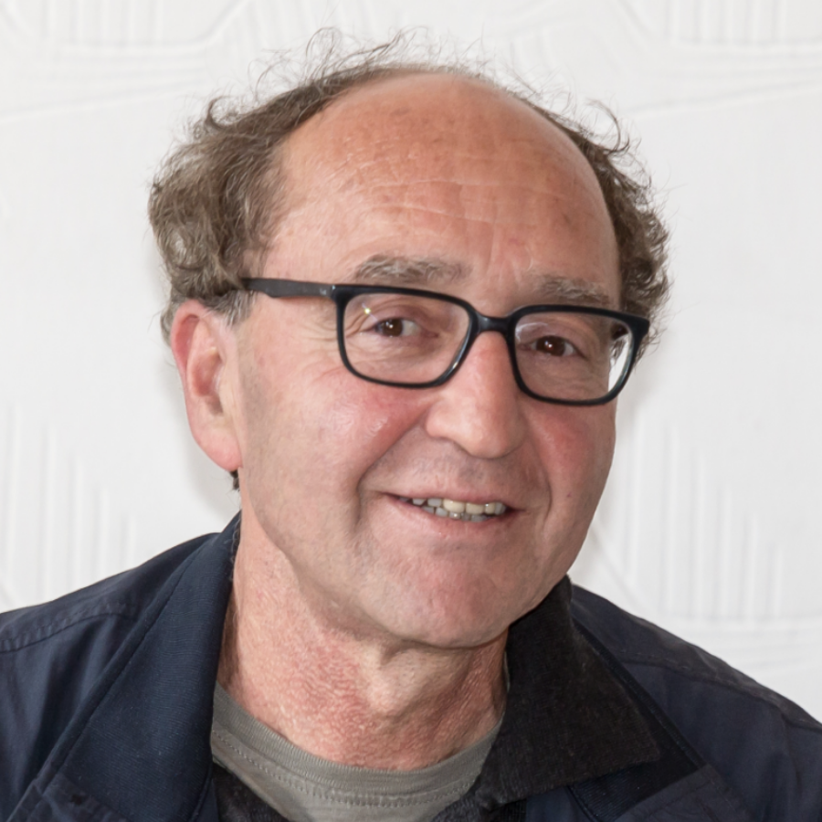 A middle-aged man with thinning hair, glasses, and a friendly smile wearing a dark jacket and gray shirt, posing in front of a white textured background.