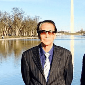A man in a pinstripe suit, blue shirt, and striped tie stands in front of a reflective body of water, with trees and a tall monument visible in the background on a sunny day.