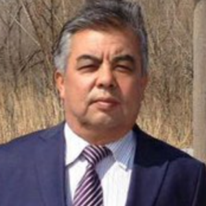 A man in a suit and striped tie stands outdoors on a sunny day, with dry grass and bare trees in the background.