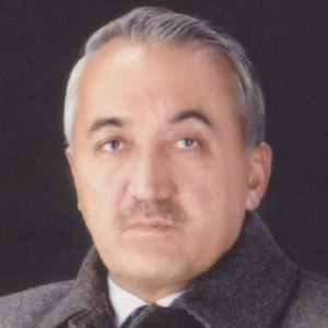A middle-aged man with short gray hair and a mustache, wearing a dark coat and a collared shirt, poses against a plain dark background.