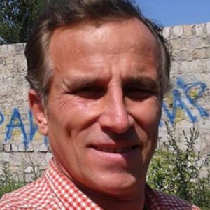 A middle-aged man wearing a red and white checkered shirt takes a selfie outdoors in front of a stone wall with blue graffiti. Trees and greenery are visible in the background under a clear sky.