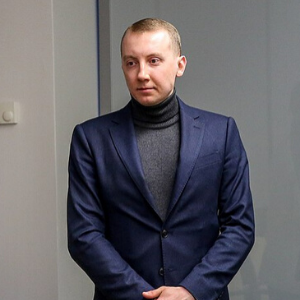 A man with short hair, wearing a navy blue suit and a gray turtleneck sweater, stands indoors with his hands clasped in front of him against a plain background.