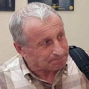 An older man with short gray hair wearing a plaid shirt looks slightly to the side. He has a backpack strap over his shoulder and appears to be in an indoor setting with blurry posters in the background.