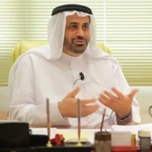 A man wearing a traditional white thobe and headscarf sits at a desk, gesturing with his hands while speaking. Office supplies and documents are visible on the desk in front of him.
