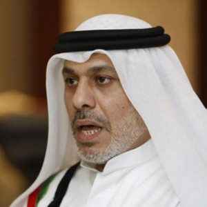 A middle-aged man with a trimmed gray beard wearing a traditional white keffiyeh and black agal, dressed in a white robe, speaking or engaged in conversation indoors.
