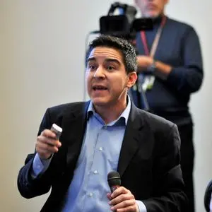 A man in a suit holds a microphone and a small device while speaking, with a cameraman filming in the background.