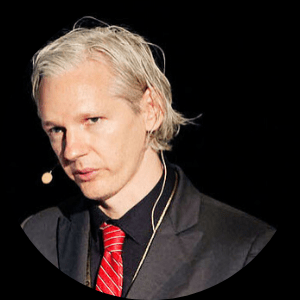 A man with pale skin and shoulder-length, light blond hair wearing a black suit, black shirt, and red striped tie. He has a headset microphone and is standing against a dark background.