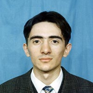 A young man with short dark hair wearing a suit, white shirt, and patterned tie poses for a formal photo against a plain blue background.