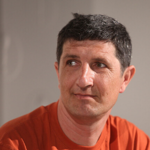 A middle-aged man with short dark hair, wearing an orange shirt, looks slightly to his left with a neutral expression. The background is plain and softly lit.