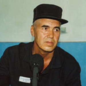 A middle-aged man wearing a black cap and dark clothing sits in front of a microphone, with a blue and white wall in the background.