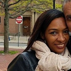 A woman with long dark hair wearing a light scarf and black jacket smiles outside near a tree and a building. A man beside her is partially visible. A red stop sign is in the background.