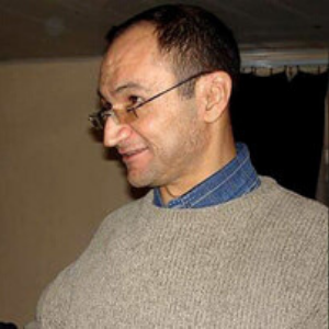 A man with short dark hair, wearing glasses, a blue collared shirt, and a beige sweater, standing indoors and smiling while facing left.