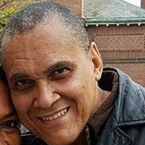 A middle-aged man with short gray hair, wearing a brown shirt and a dark jacket, smiling at the camera. A partial view of another person is visible on the left side. Brick building in the background.