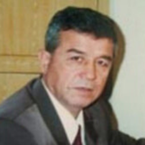 A middle-aged man with short dark hair wearing a suit and tie is sitting indoors, looking slightly past the camera. There is a wooden door and light-colored wall in the background.