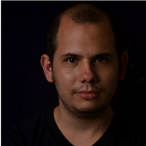 A man with short hair and a trimmed beard is looking directly at the camera against a dark background. He is wearing a black shirt and has a neutral expression.