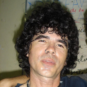 A person with curly black hair and a nose ring is looking at the camera. They are indoors, standing in front of a wall with handwritten notes or drawings in the background.