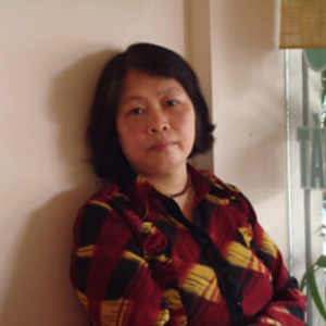 A woman with short dark hair wearing a red, yellow, and black plaid shirt stands indoors, leaning against a light-colored wall, looking at the camera with a neutral expression.