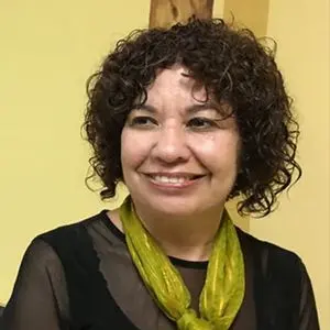 A woman with short, curly brown hair smiles while looking slightly to the side. She wears a sheer black top and a bright green scarf against a yellow background.