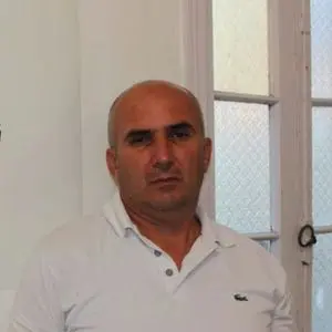 A man with a shaved head wearing a white polo shirt stands indoors in front of a window with frosted glass panels.