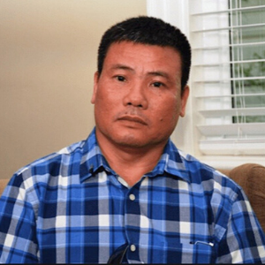 A man with short black hair wearing a blue plaid shirt sits indoors on a beige couch, looking directly at the camera. White blinds and a cream-colored wall are in the background.