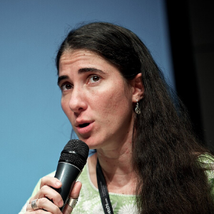 A woman with long dark hair speaks into a microphone. She wears a green top, dangling earrings, and a lanyard, and appears to be addressing an audience. The background is plain and softly lit.