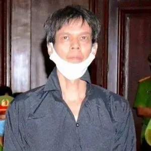 A man with short dark hair wearing a black shirt and a white face mask pulled below his chin stands indoors in front of wooden doors, with blurred people in green uniforms in the background.