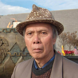 An older man wearing a patterned hat and brown blazer stands outdoors near a monument, with small flags and flowers visible in the background on a partly cloudy day.