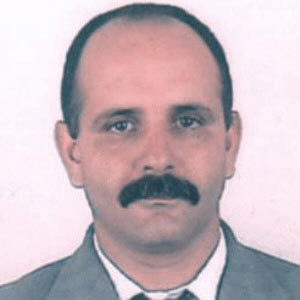 Portrait photo of a man with short dark hair, a mustache, and wearing a grey suit jacket, white shirt, and tie, against a plain light-colored background.