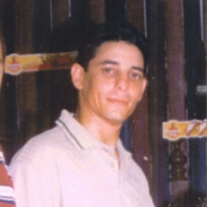 A young man with short dark hair wearing a light-colored collared shirt stands indoors in front of a wooden background with colorful stickers or signs.