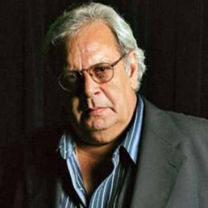 An older man with gray hair and glasses wearing a dark suit jacket and a striped shirt stands against a dark background, looking seriously at the camera.