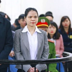 A woman in a gray blazer and white shirt stands in a courtroom, flanked by people in uniform and others seated in the background.