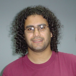 A person with curly black hair, wearing glasses and a maroon t-shirt, is smiling slightly in front of a plain gray background.