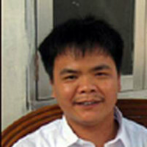 A man with short black hair and a mustache is smiling, sitting in front of a window. He is wearing a white collared shirt and is seated on a wooden chair.