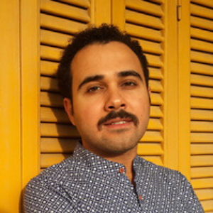 A man with short curly hair and a mustache, wearing a navy blue patterned shirt, stands in front of yellow wooden shutters, looking at the camera with a slight smile.